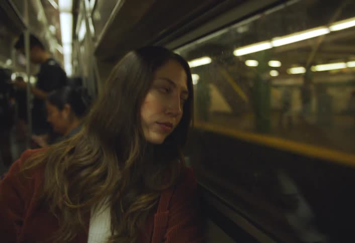 A young woman in a rustcolored jacket gazes out the window of a moving subway train as other passengers sit and stand around her