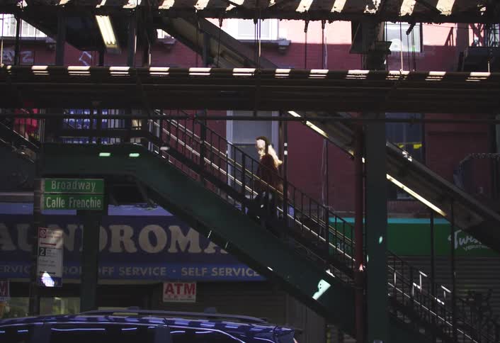 Two women and a man are each walking separately up the stairs of an elevated train station on a sunny day as sunlight reflects off a building beside the staircase