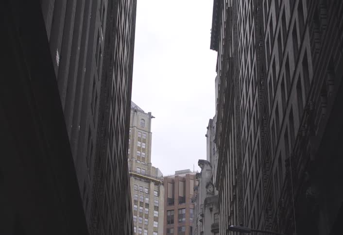 A narrow view looking up between tall city buildings reveals a cloudy sky and the tops of other urban skyscrapers in the background