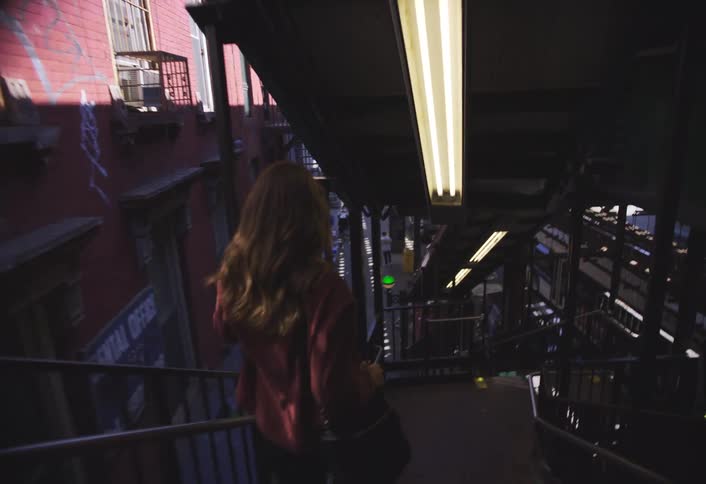 A woman walks down a stairway from an elevated platform in an urban area and exits onto a busy city sidewalk with storefronts