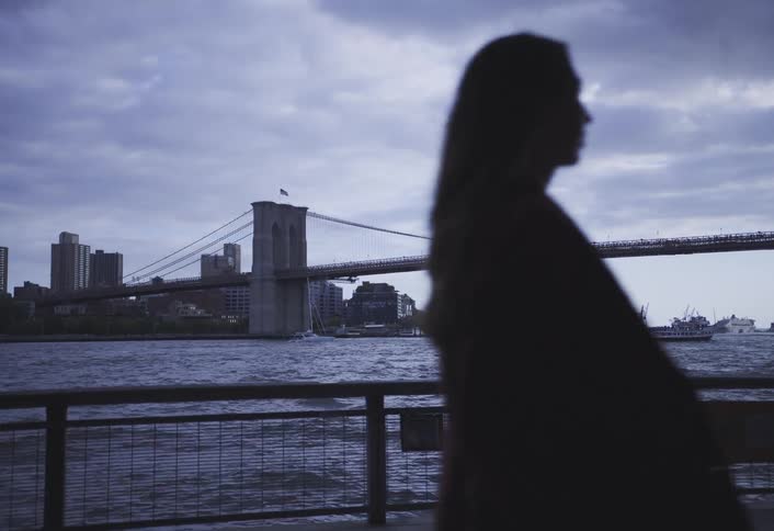 Silhouetted woman walks past waterfront railing with brooklyn bridge and city skyline in the background at dusk