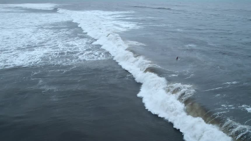 An aerial view captures a person on a surfboard in the ocean near a large breaking wave under an overcast sky