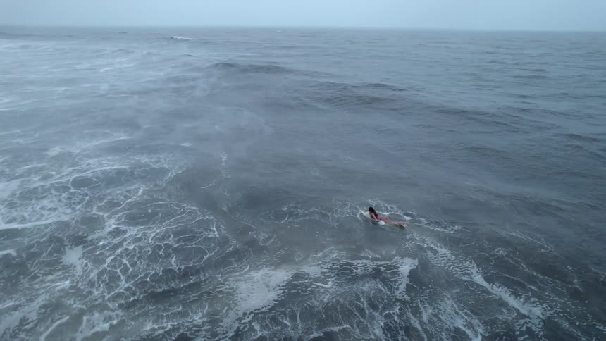 An aerial view shows a lone surfer paddling through rough choppy waters on a gloomy day
