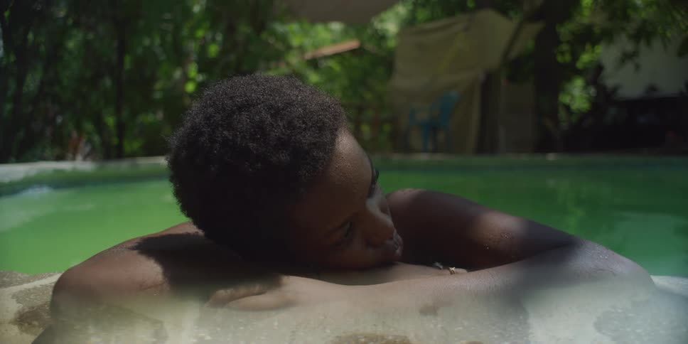 A person with short hair rests their head on the edge of a swimming pool with lush greenery visible in the background