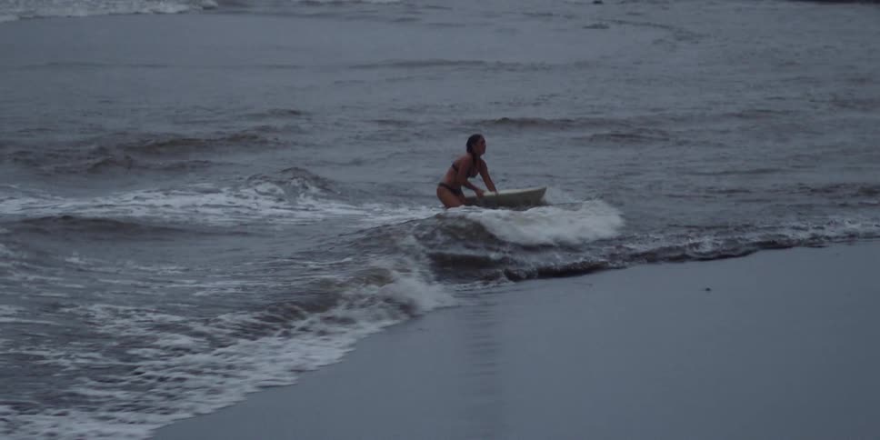 Woman stands and walks ashore with surfboard as waves crash over her