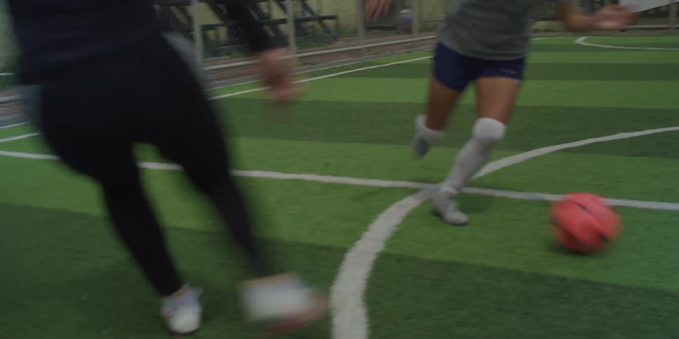 Soccer players compete intensely on a green artificial turf field with one kicking a vivid orange ball