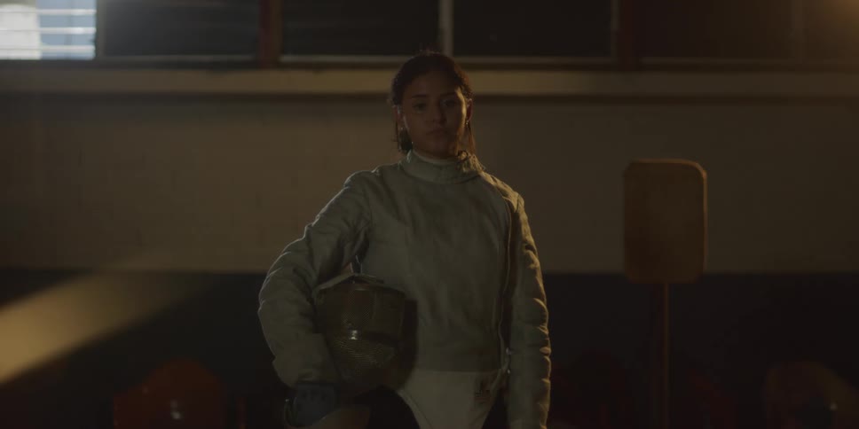 A fencer in full gear holds a fencing helmet under one arm standing in a dimly lit room with a wooden target in the background