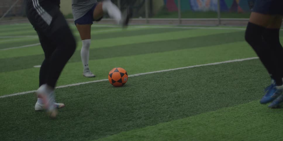 Three individuals are stretching and preparing on a green turf soccer field near an orange soccer ball