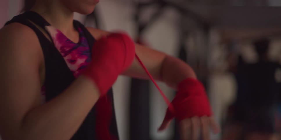 A woman with her hair pulled back wraps her hands with red boxing wraps her expression focused and serious