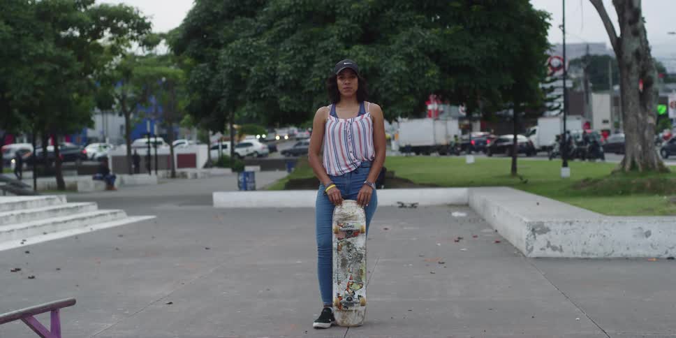 A woman stands on a concrete surface in a park holding a skateboard vertically with trees parked cars and a busy street in the background