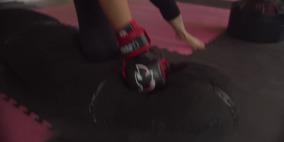 A woman wearing gloves and gym pants barefoot aggressively punches a punching bag on the floor at midday