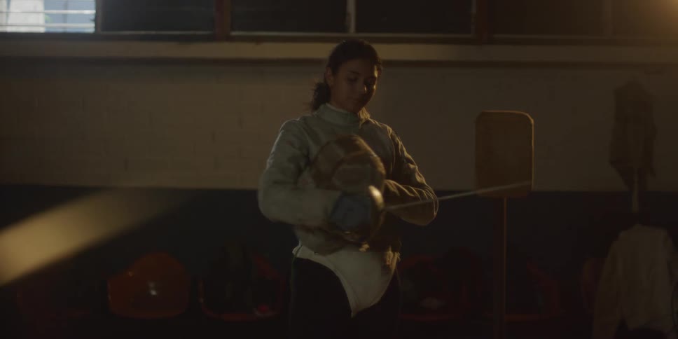 Female fencer removes her fencing mask and stands indoors while holding her epee