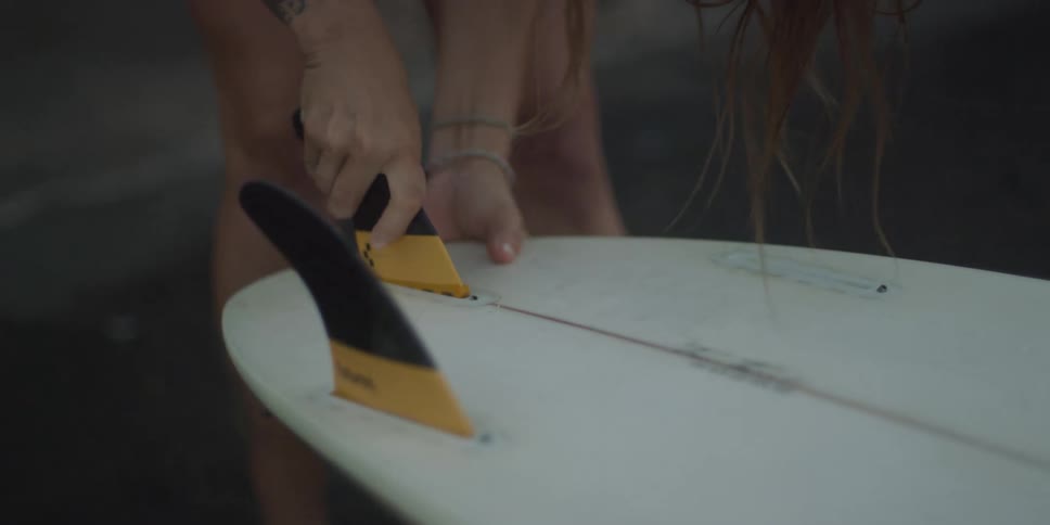 A blonde woman secures a fin onto a surfboard with her hands