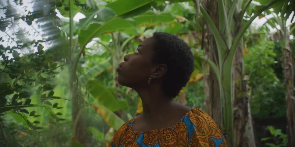 A young black woman in a colorful patterned top gazes upwards amidst a lush tropical environment filled with banana trees