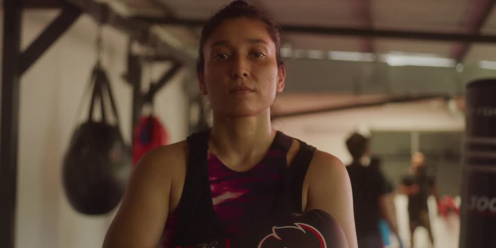 A woman with boxing gloves is standing near a punching bag while men walk around behind her