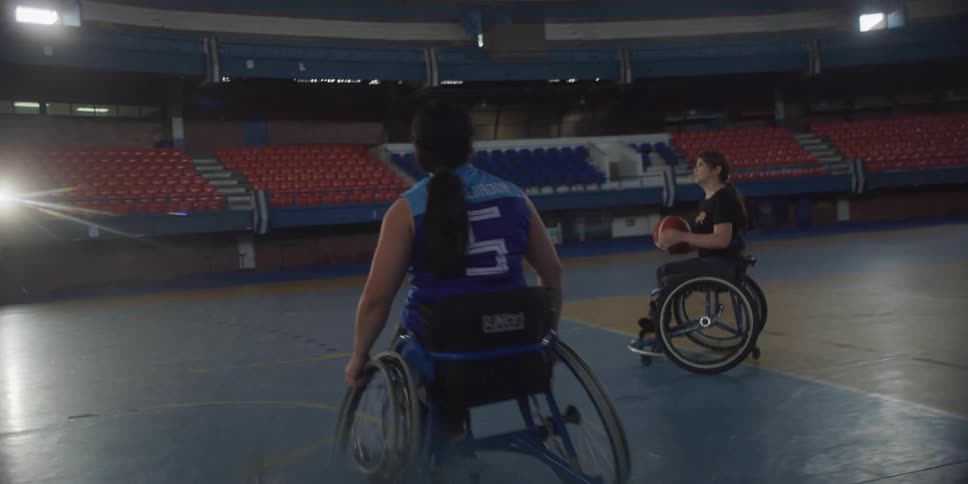 Two disabled women in wheelchairs are playing basketball passing the ball to each other on a basketball court as sunlight shines through several openings in the stadium