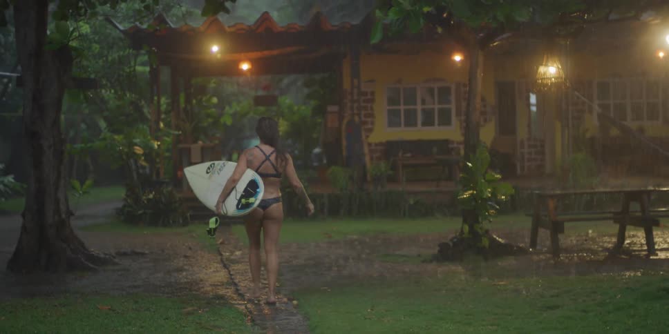Woman in bikini walking in the rain with a surfboard towards a house