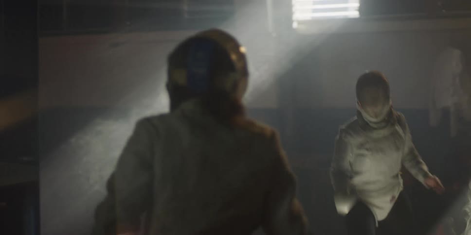 Two fencers in protective gear and helmets face each other in a dimly lit smoky arena