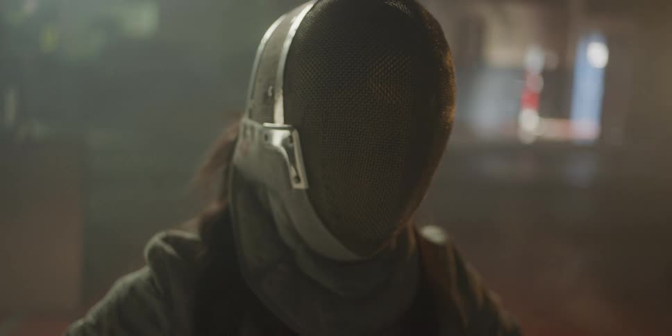 A woman reaches up with her right hand and pulls down a fencing helmet covering her face