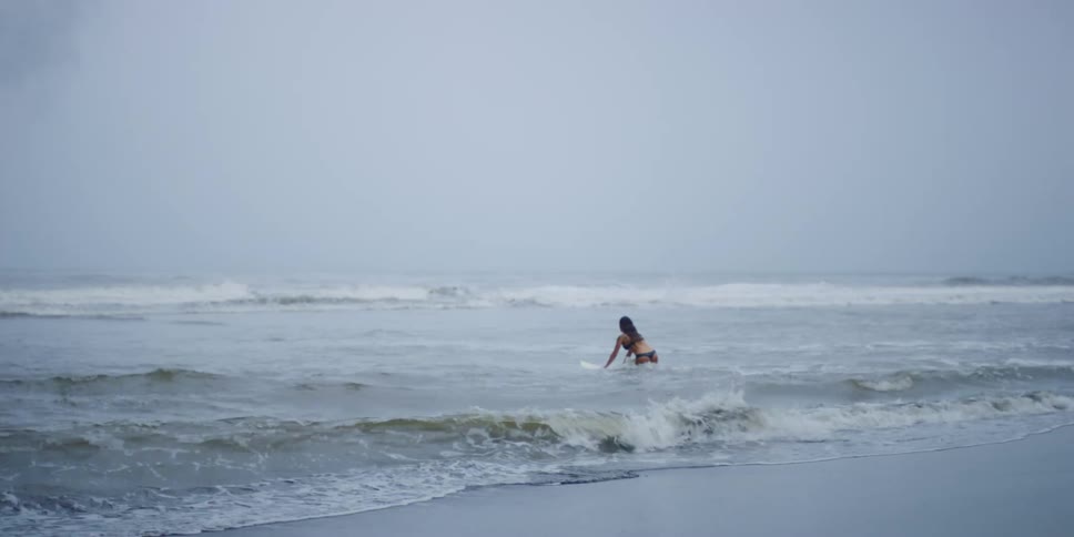 Woman in bikini wading into rough sea with surfboard on cloudy day