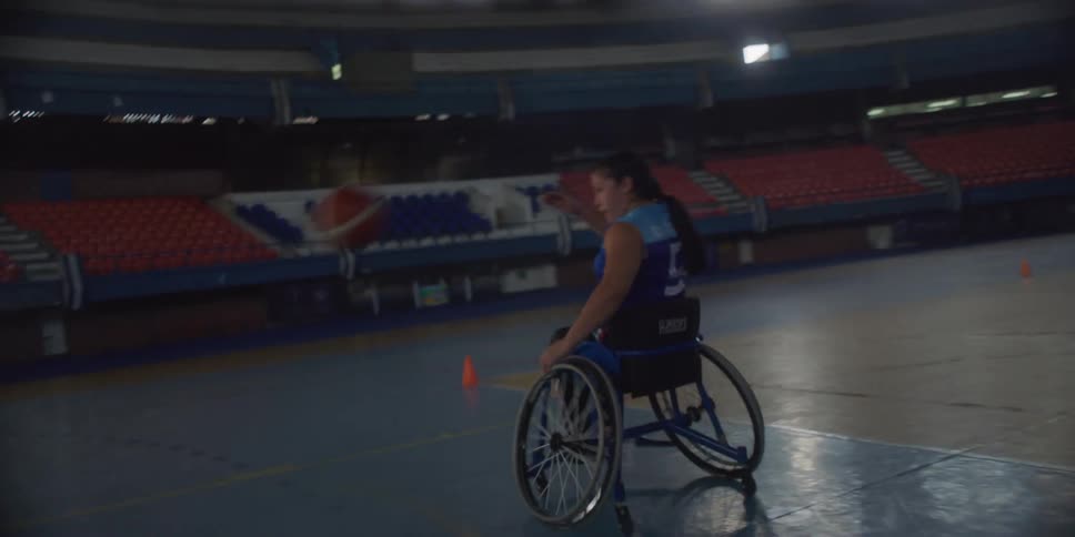 Two disabled women in wheelchairs are playing basketball passing the ball to each other on a basketball court as sunlight shines through several openings in the stadium