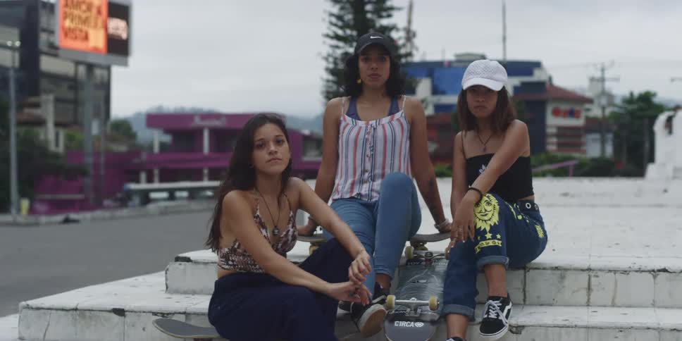 Three teenage girls are sitting on white steps holding skateboards surrounded by urban buildings and leafy trees