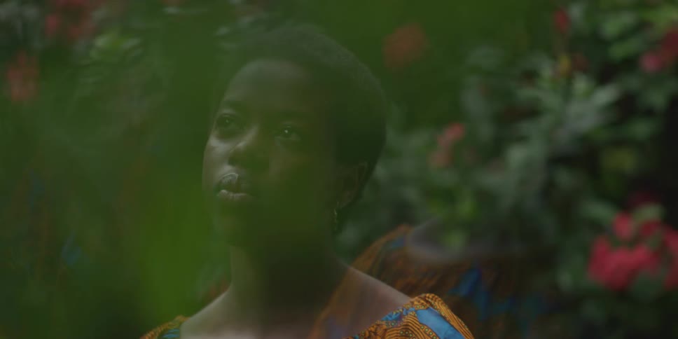 A young black woman with short hair wearing a colorful patterned dress looks pensively off to the side in a garden setting