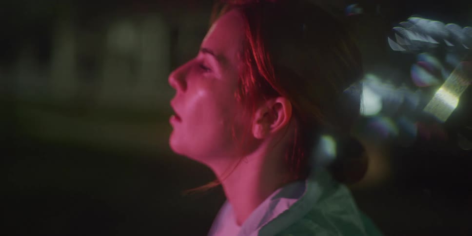 Bathed in red light a woman with a determined expression faces forward tears welling in her eyes against a blurred urban background with bokeh effects