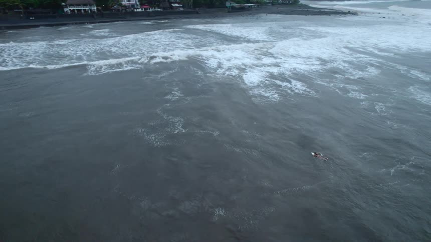 A lone surfer awaits a wave on a dark choppy sea near a shore with buildings