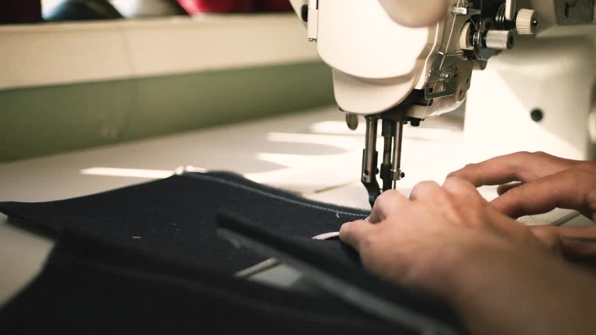 A person operates a sewing machine carefully guiding fabric with their hands surrounded by spools of thread