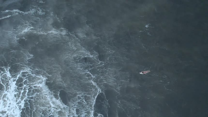 Turbulent ocean waves crash around a lone surfer paddling on a surfboard