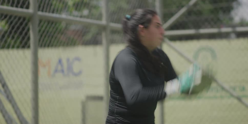 A female goalkeeper dives to catch a football on a field surrounded by a wire fence