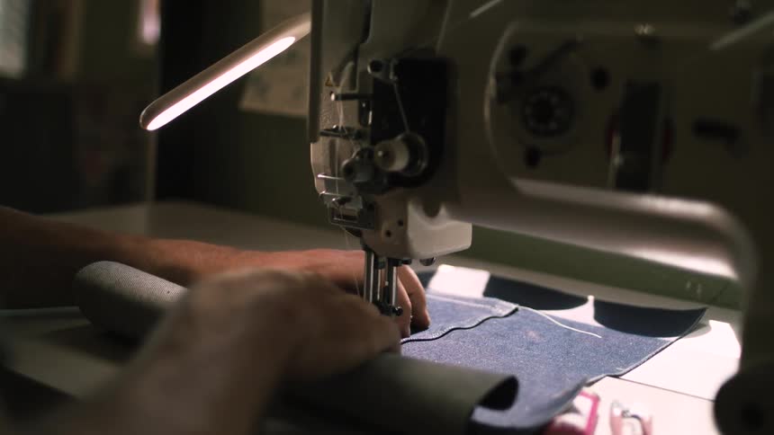 Man sews two fabric pieces together using a sewing machine while a table lamp illuminates his task
