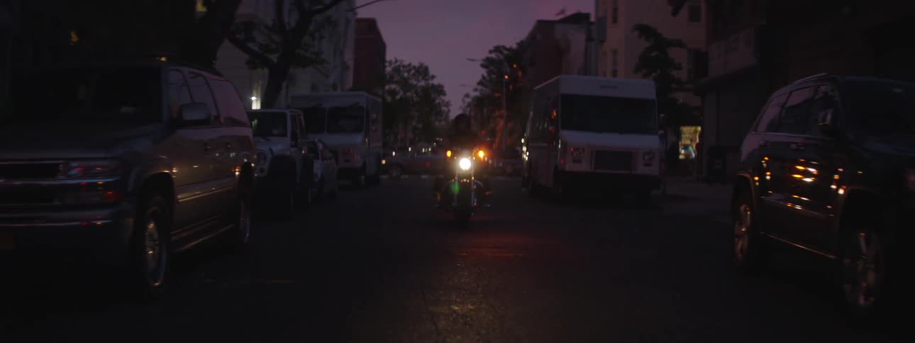 A bike rides along the street at dusk with cars and a van parked on both sides of the road