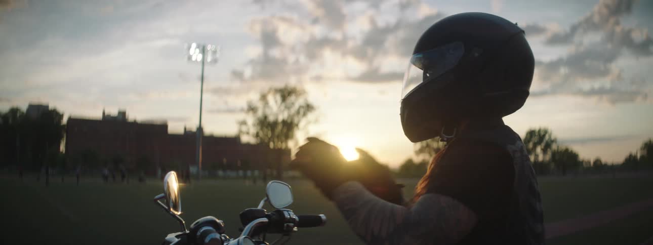 Motorcyclist pulls up to the edge of a school sports field occupied by student athletes at sunset