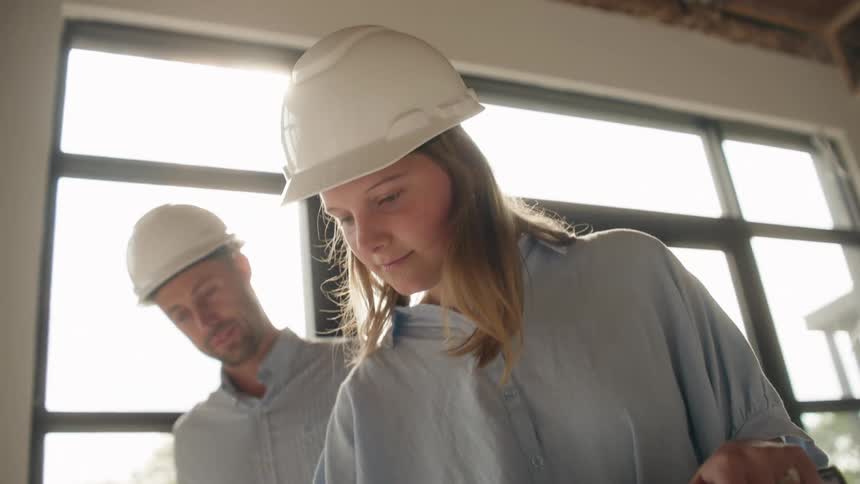 Two construction employees are flipping through pages of blueprint plans while standing indoors