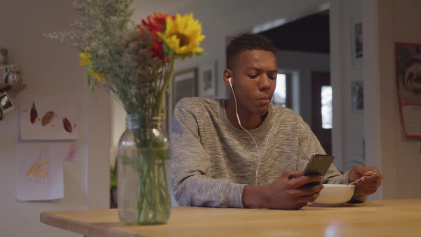A guy eats a bowl of cereal while on his phone