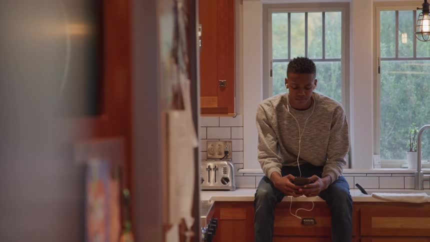 A young black teenager sits on a kitchen countertop at midday wearing wired earbuds and smiling while using his phone