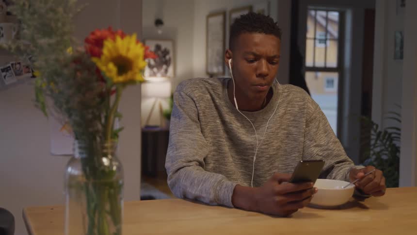 A teenage boy sits at the dining table eating wearing white wired earbuds and smiling at his phone with a flower plant in a glass vase next to him