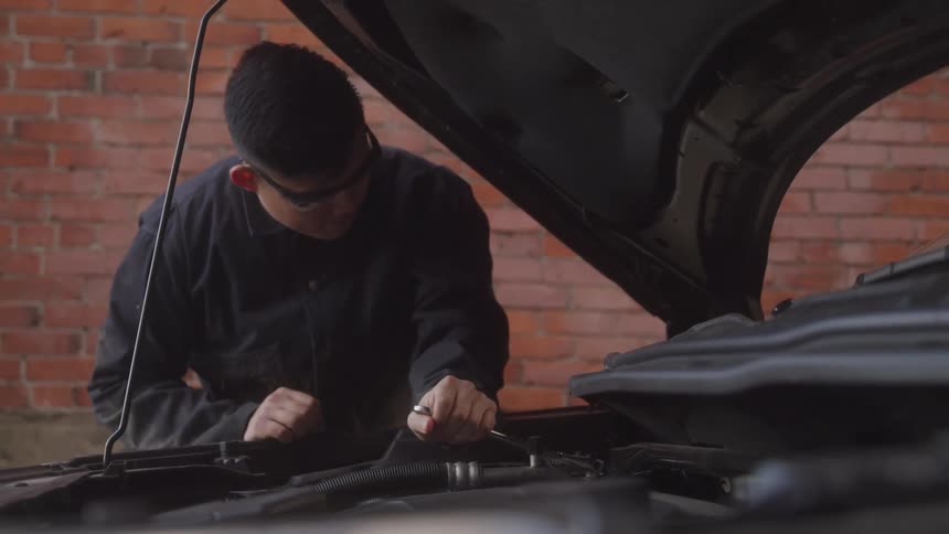 Mechanic working on a car engine with the hood open wearing protective glasses and a dark uniform