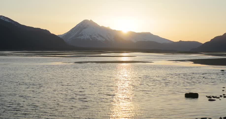 The sun sets behind a snowcovered mountain near a shallow lake bed