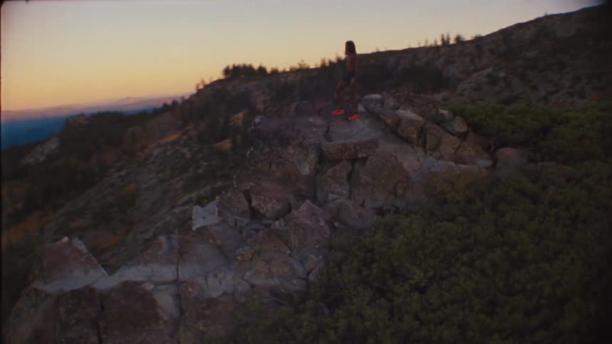 A person stands on a rocky peak overlooking a mountain landscape at sunset