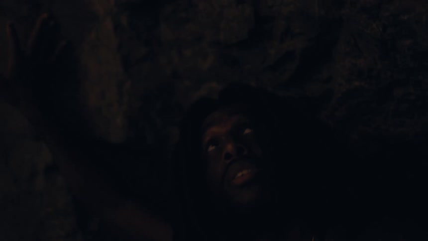 The face of a man with long hair who is coming out of a rocky cave and looking up
