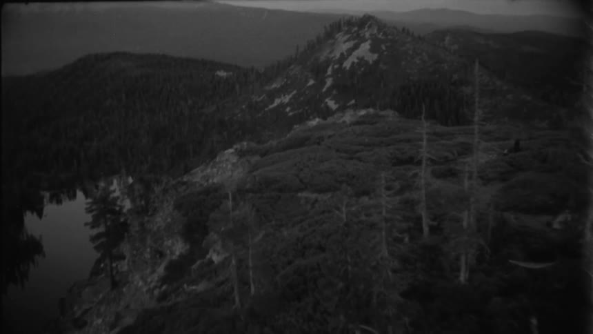 A man stands at the edge of a cliff with a view of a mountain stream and a forest stretching out below under the midday sun