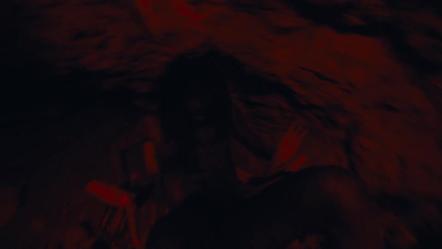 A caveman sits on the floor inside a cave and begins to rise as someone approaches