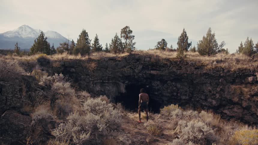 Shirtless man jogs toward the hillside cliff with snowcapped mountains in the distance