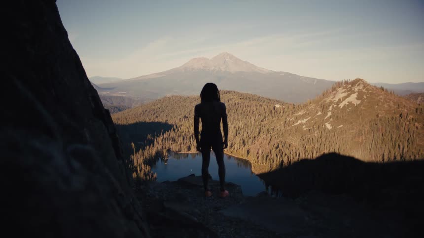The silhouette of a man standing on top of a rock on a mountain looking at a landscape of forest and pine trees with a huge mountain in the background