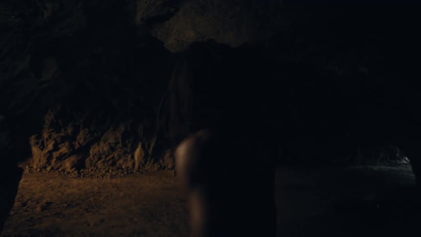 A shirtless hairy man is standing in a rocky cave and then he starts walking into it