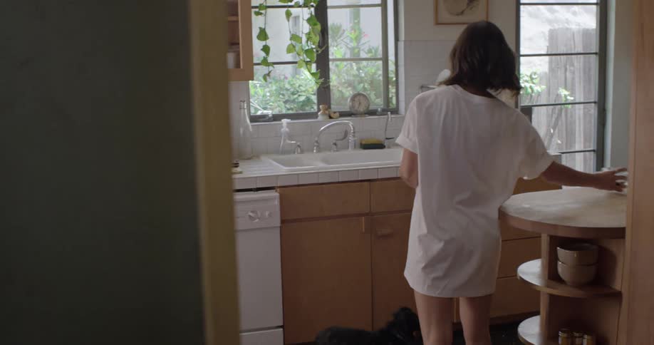 A young woman kneels petting her dog before walking to the kitchen sink with a cup
