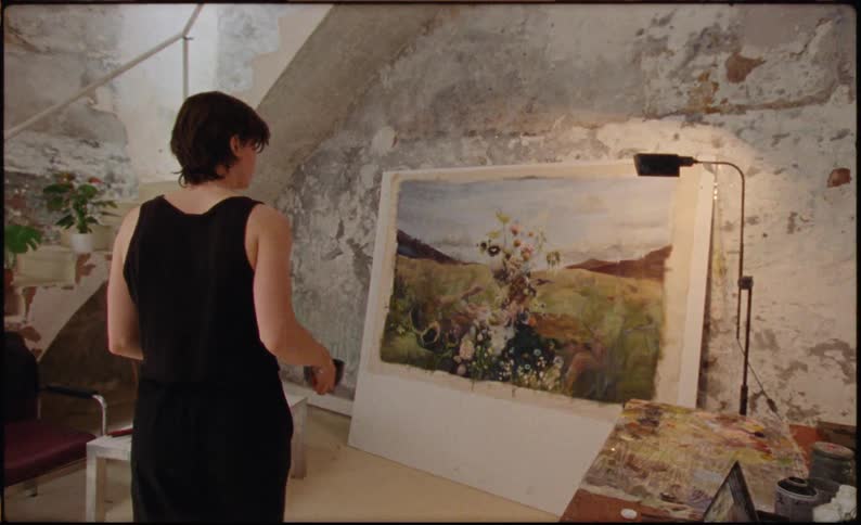 Artist picking up a pot and looking at a painting in a frame